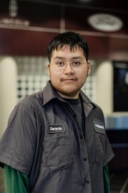 Man with glasses, wearing a black jacket, standing in a building with dark-colored walls.