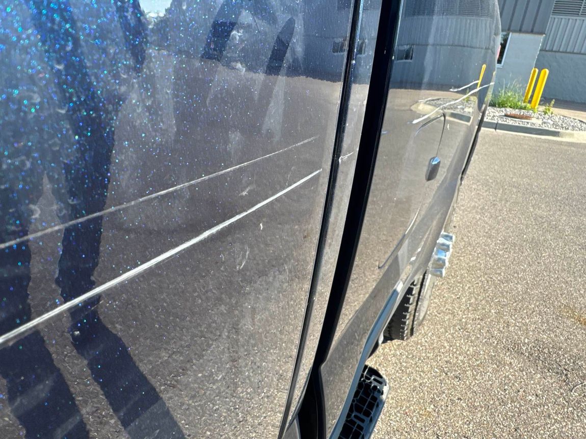 Close-up of a dark-colored vehicle's side with scratches and dirt. Bright sun illuminates the surface.