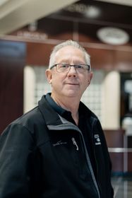 Man with glasses, wearing a black jacket, standing in a building with dark-colored walls.