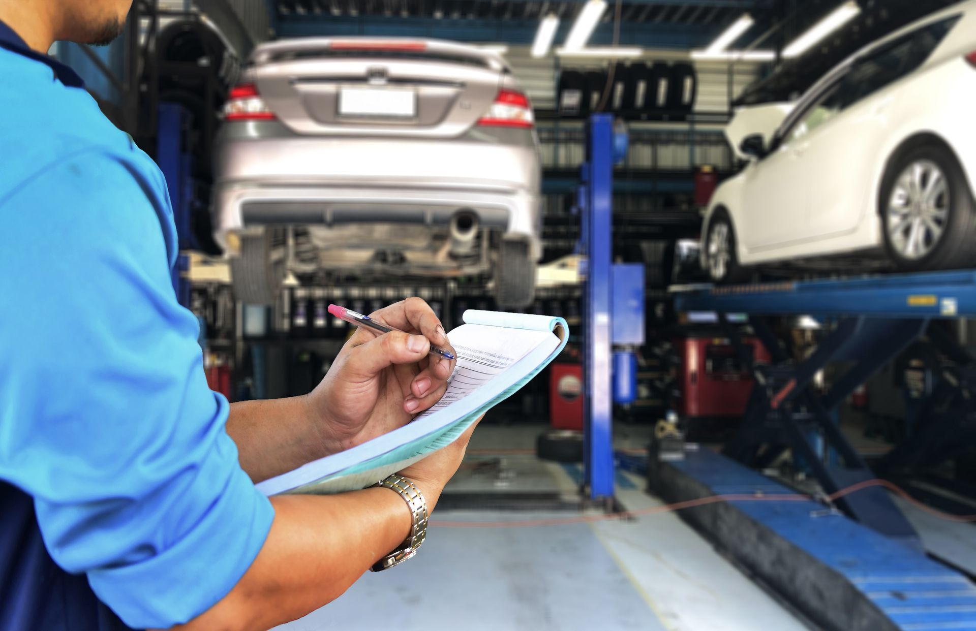 Mechanic in blue shirt taking notes while inspecting a car on a lift in a garage.