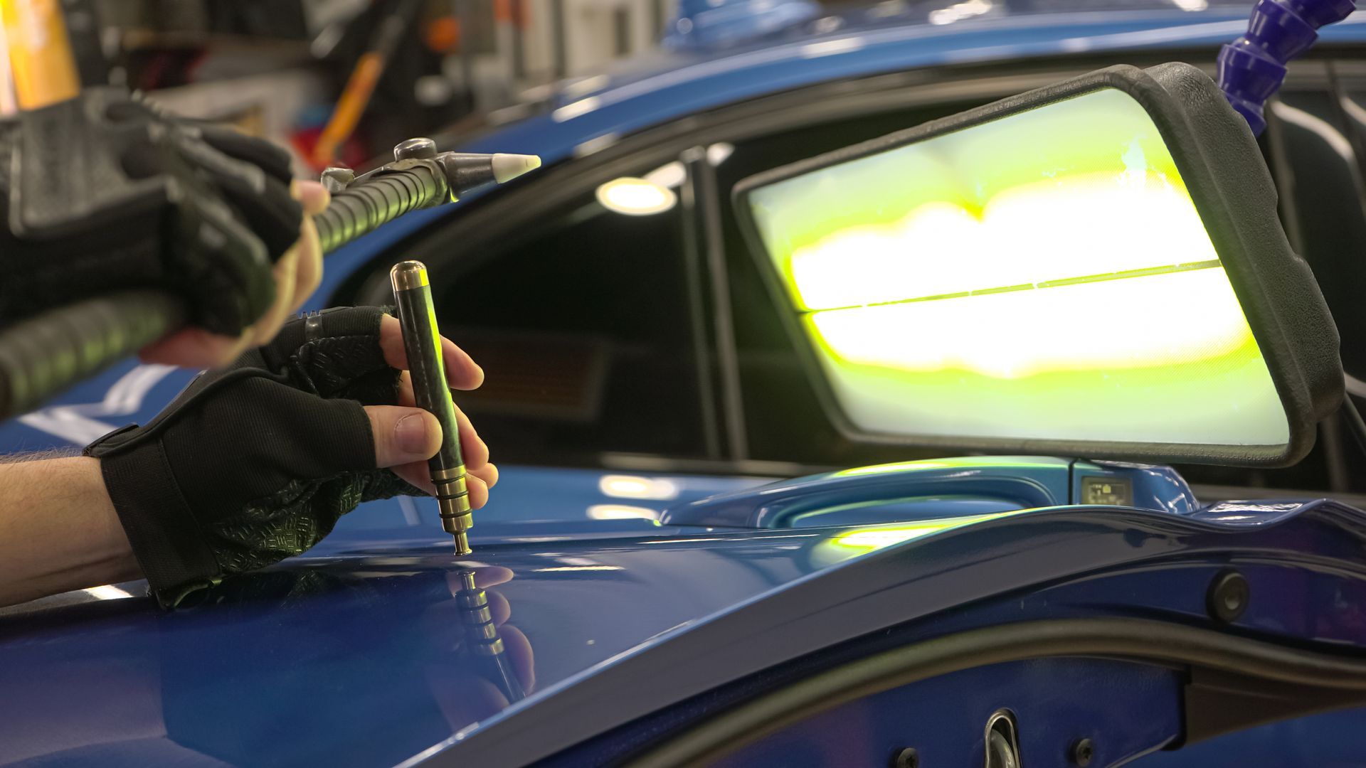 A person uses a tool to remove a dent from a blue car's hood in a repair shop, aided by a reflective light.