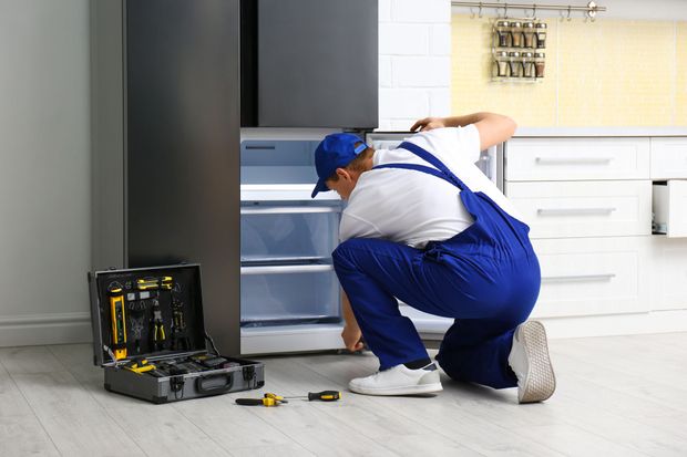 A repairman in blue overalls kneels fixing a refrigerator, tools in an open toolbox.