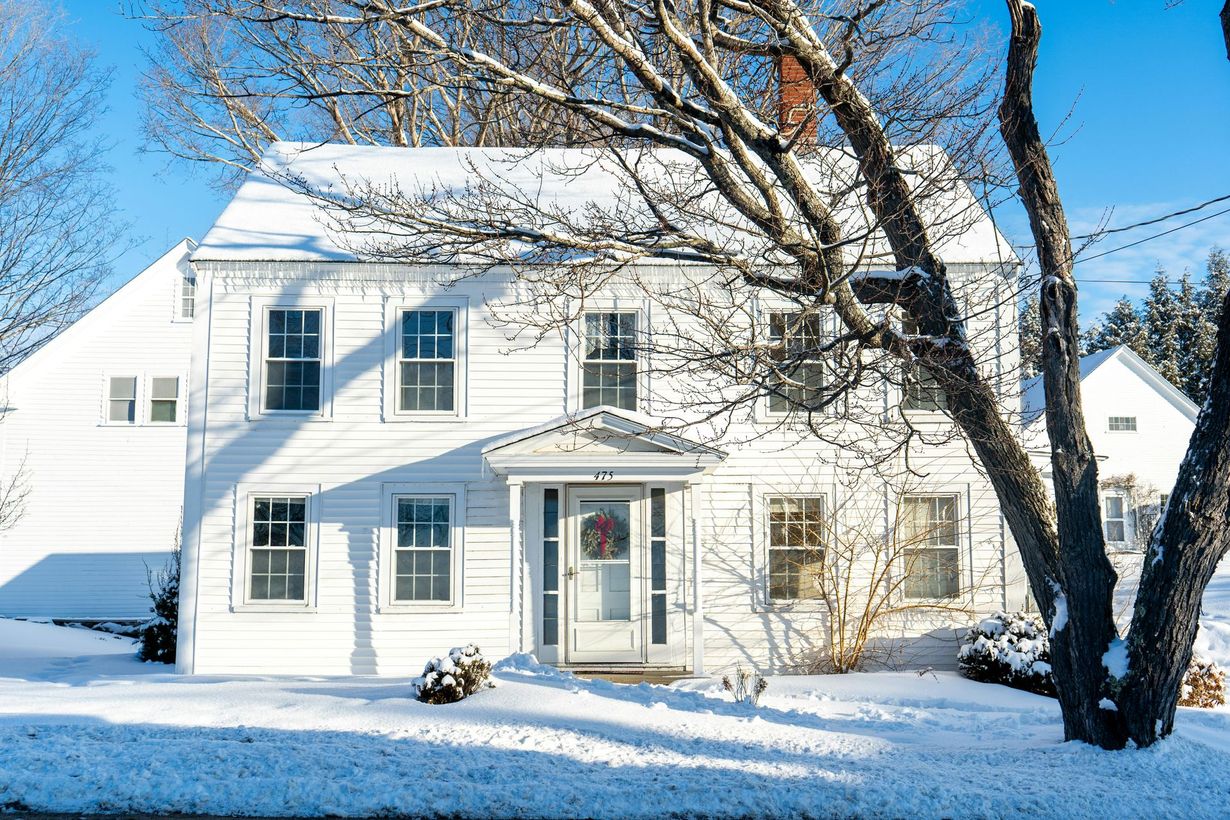 A two-story white house with a snow-covered yard, sunny sky, and a large tree in the foreground.