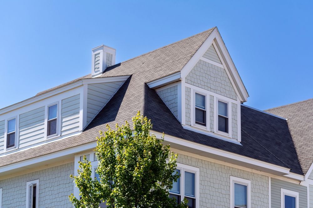 A light blue house with cedar shingle siding, a dark grey shingled roof, and a prominent dormer window under a blue sky.