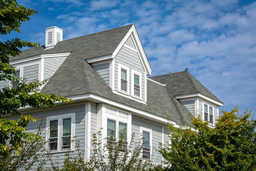 A multi-story grey house with white trim and a shingled roof under a bright blue sky with scattered clouds.