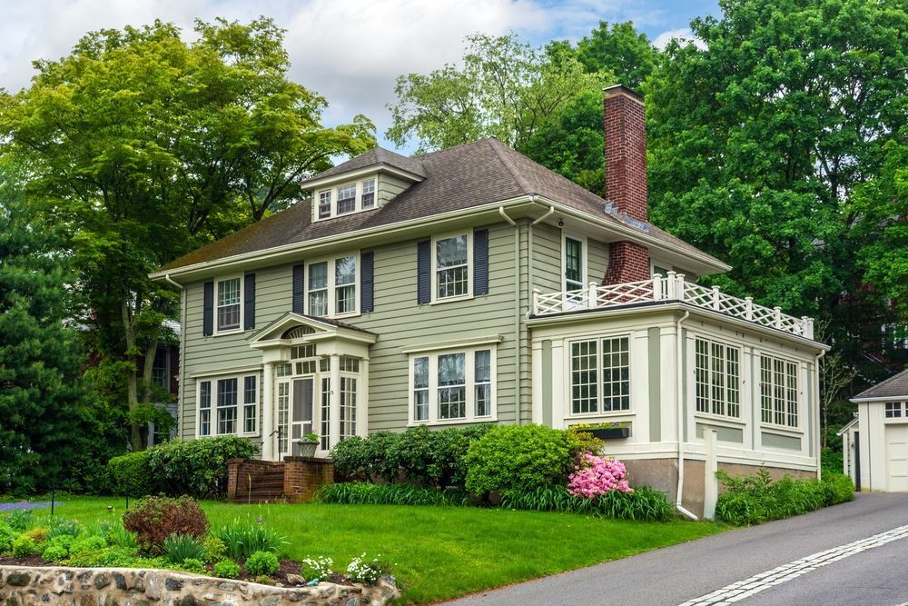 A light green two-story house with a brick chimney and a side porch, surrounded by lush green trees and landscaping.