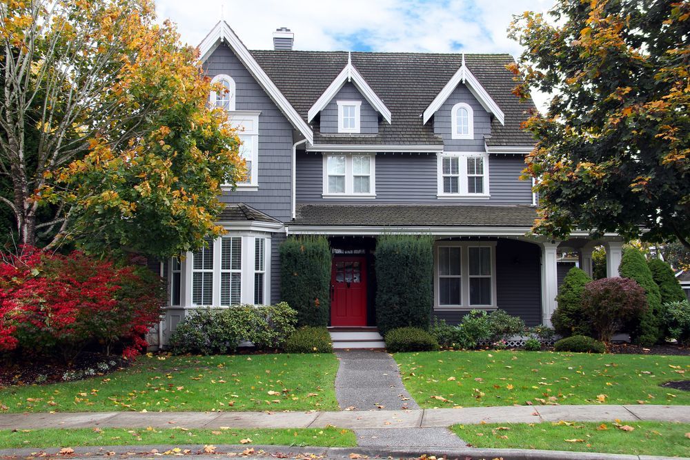 A two-story grey craftsman house with three dormers, a bright red front door, and a front lawn with trees.