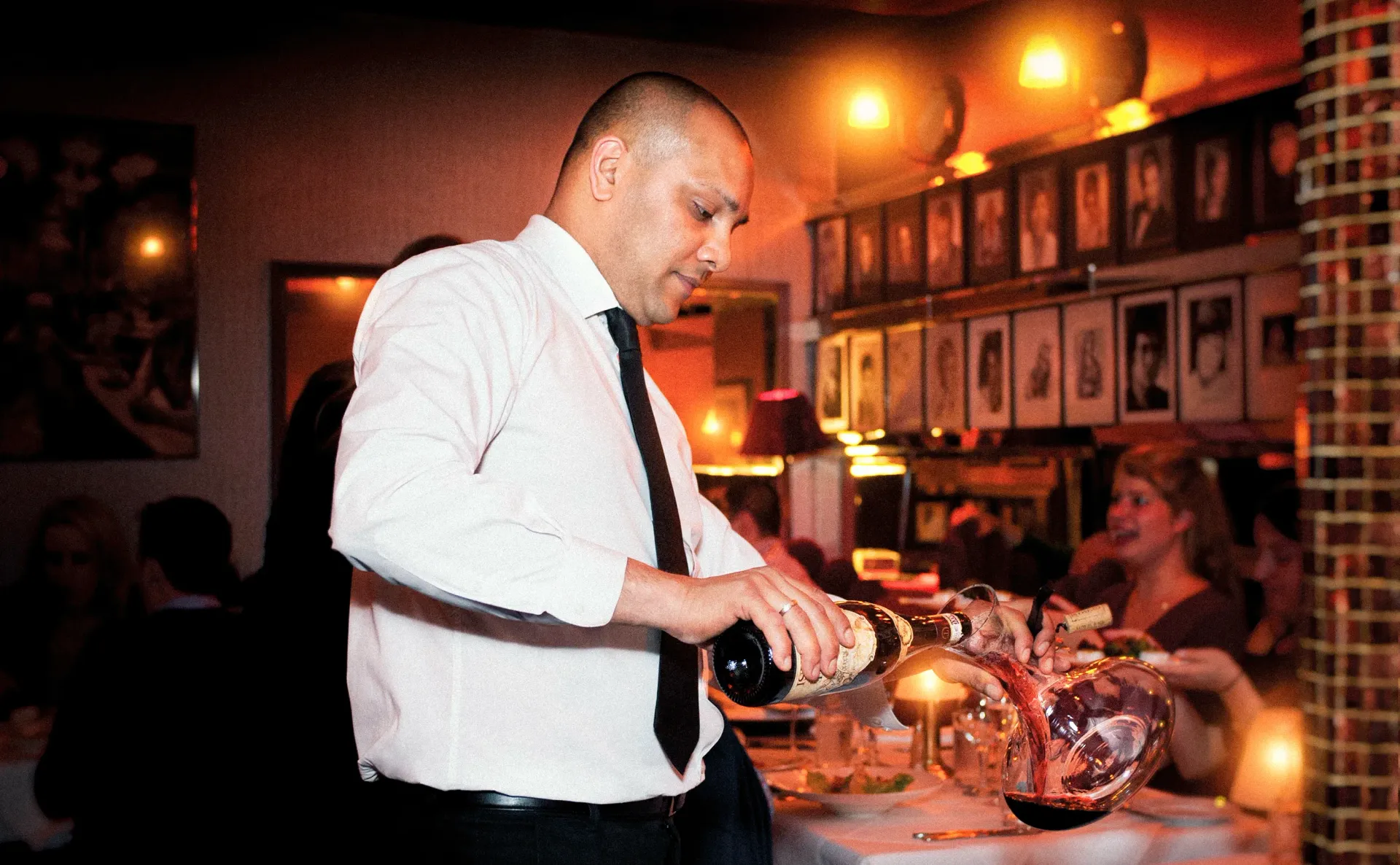 Server pouring wine at a restaurant; red lighting, photos on wall.