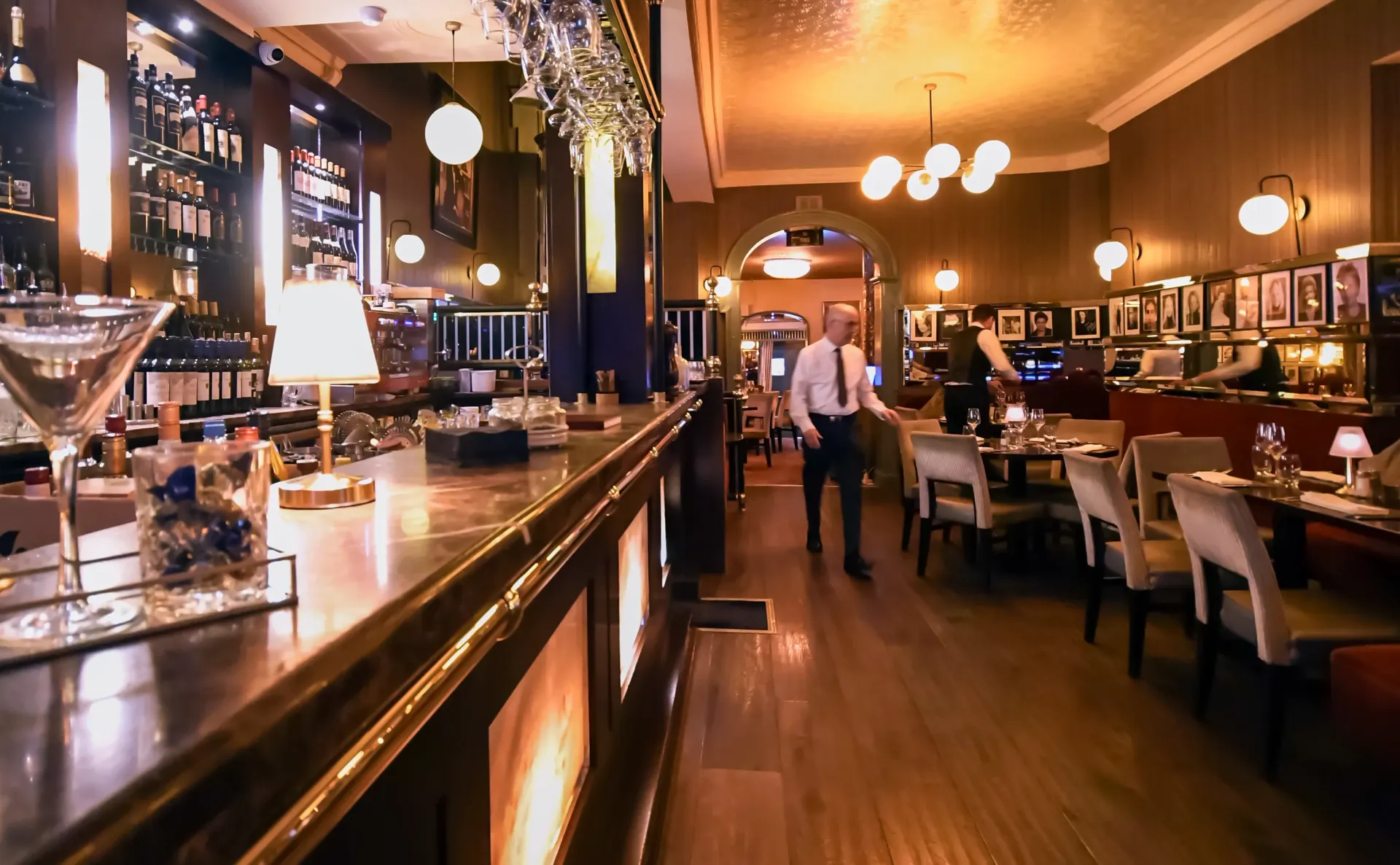Interior of a bar and restaurant with a long bar, staff, and tables with customers. Warm lighting and dark wood.
