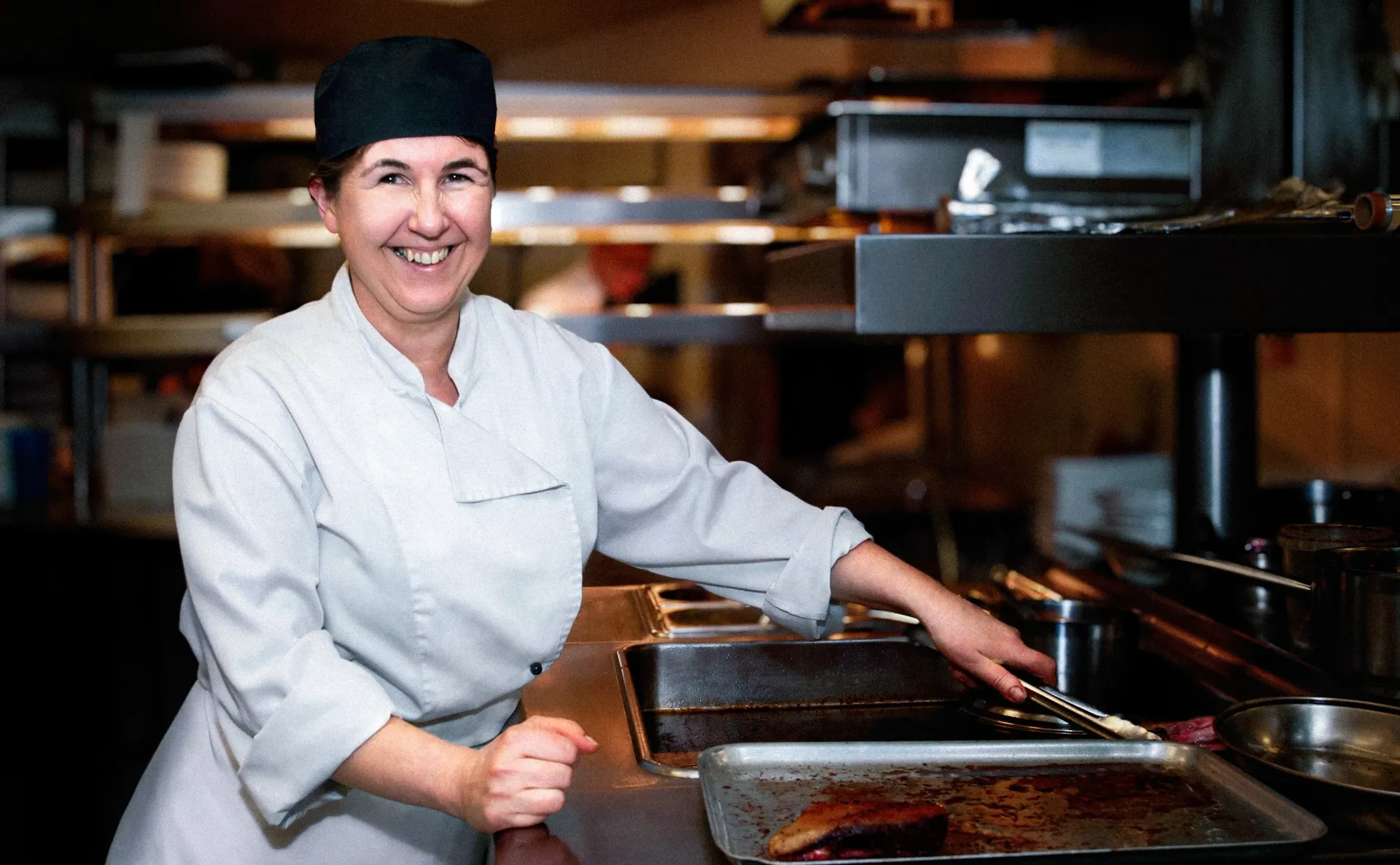 Chef in white uniform and black hat, smiling while working in a restaurant kitchen.