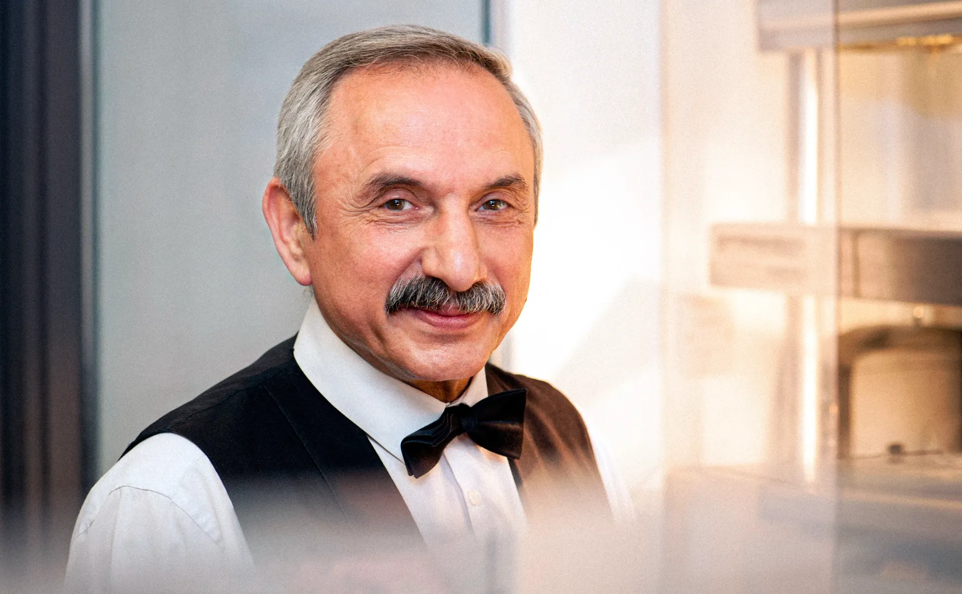 Man with mustache and bow tie, wearing a vest and white shirt, smiling.