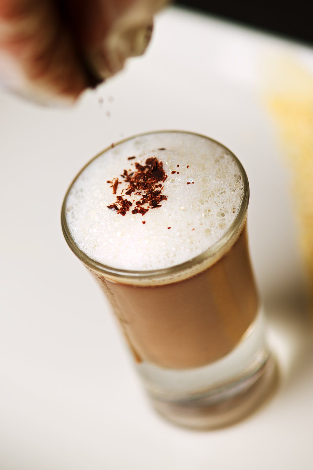 A cappuccino in a glass, being sprinkled with cocoa powder, on a white surface.