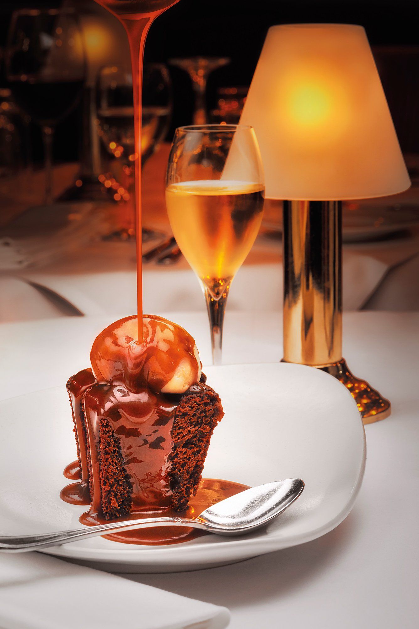 Molten chocolate cake with ice cream, caramel sauce drizzled, on white plate, beside a glass and lamp.