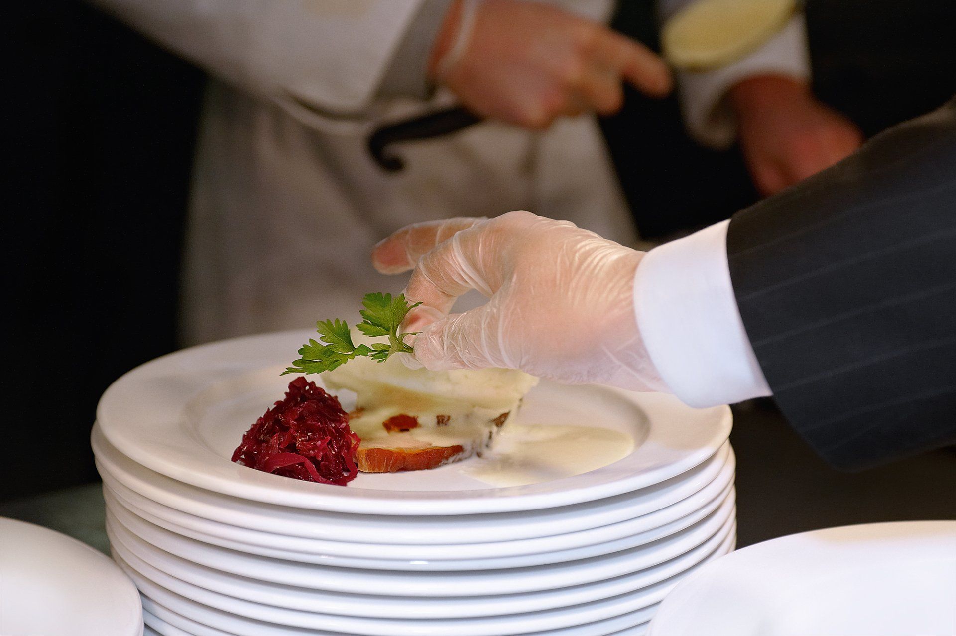 Chef plates a dish with garnish; other chef adds sauce.