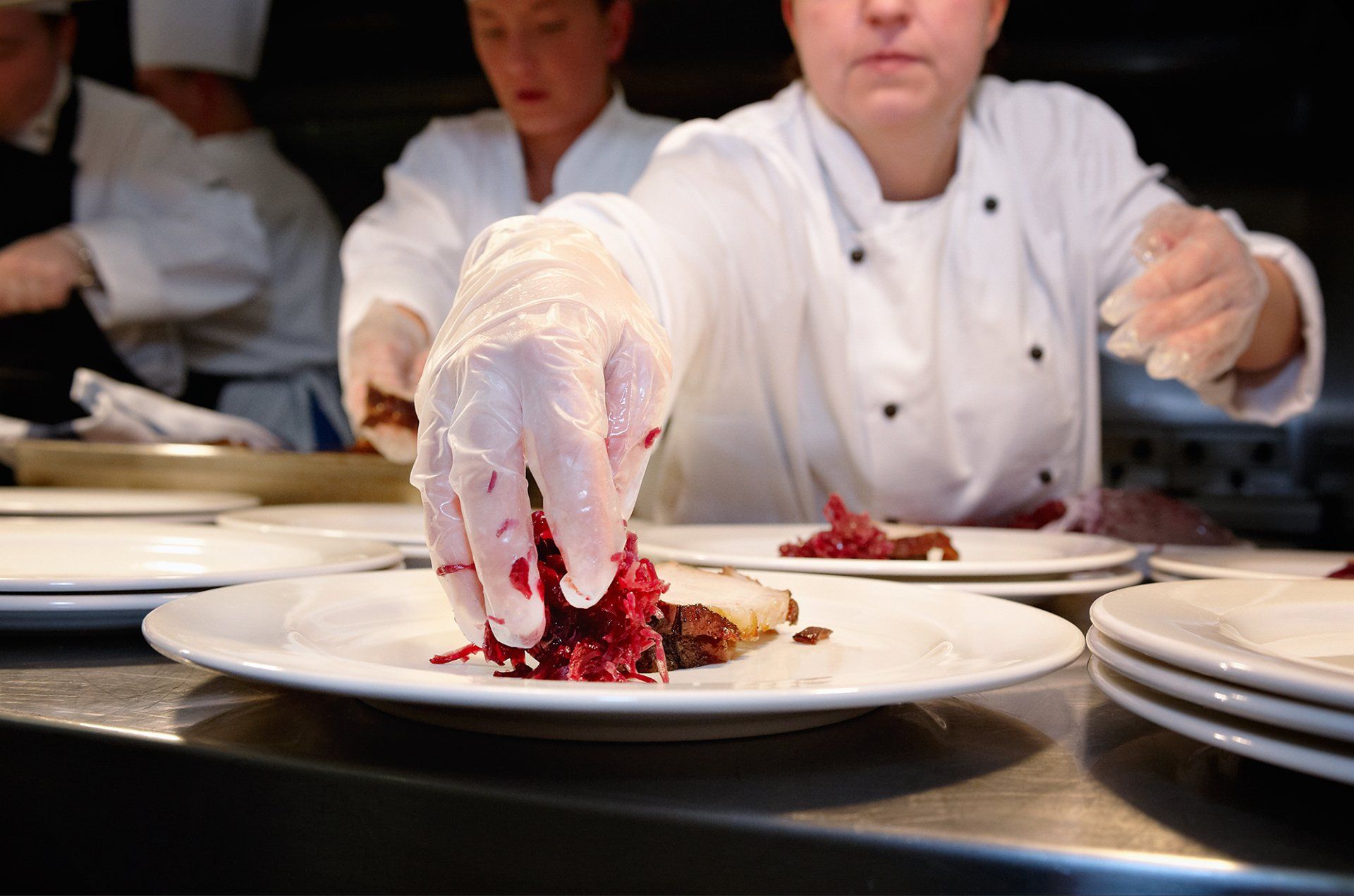 Chef plating food, placing red cabbage on a plate. Kitchen setting with other staff.