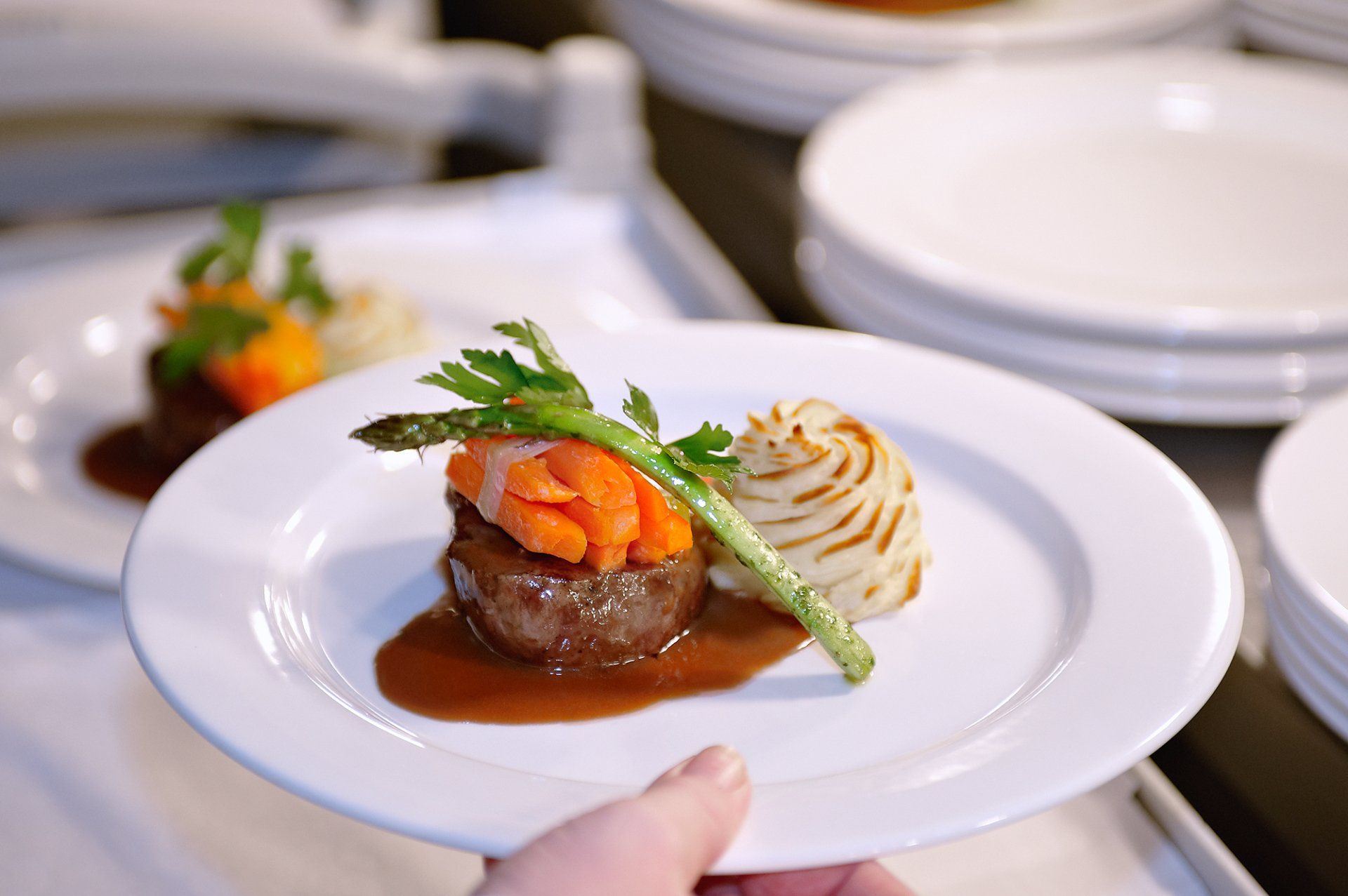 A hand holding a plate with steak, mashed potatoes, asparagus, and carrot garnish, drizzled in sauce.