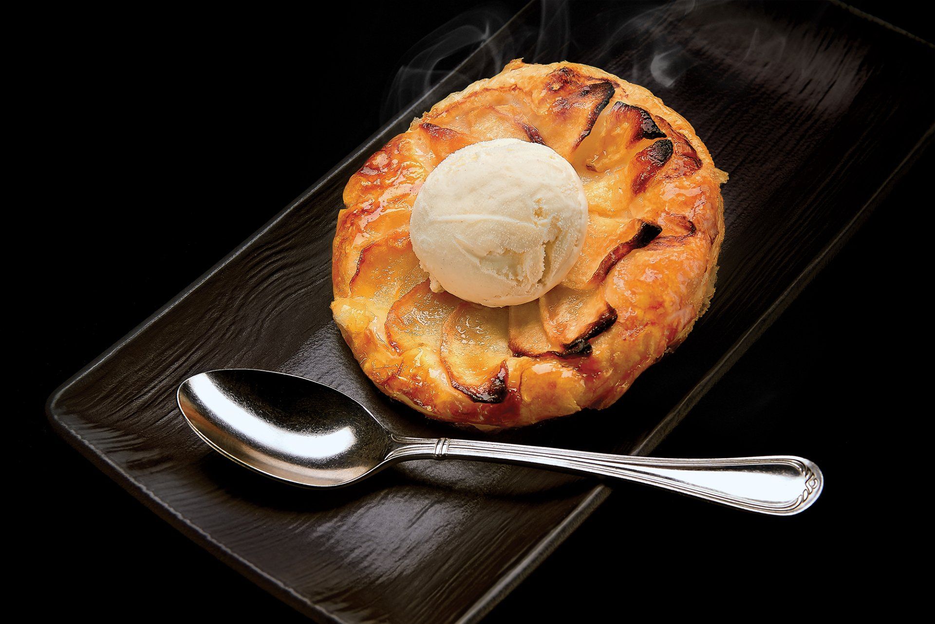 Apple tart with vanilla ice cream and spoon on a black serving platter.