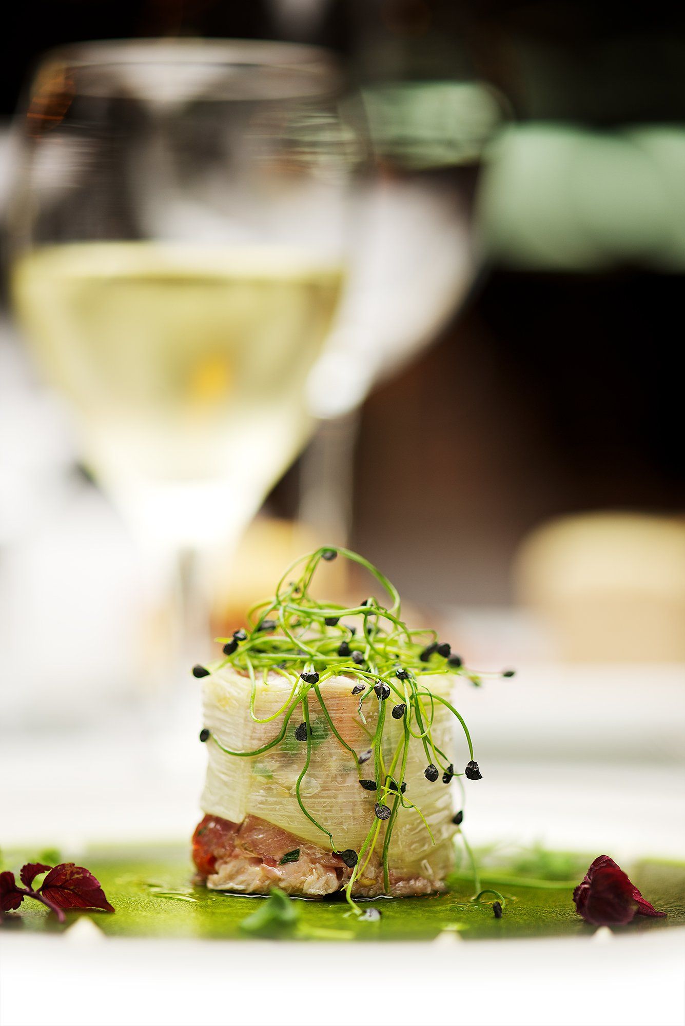 A stack of food garnished with sprouts, on a green sauce, in front of wine glasses.