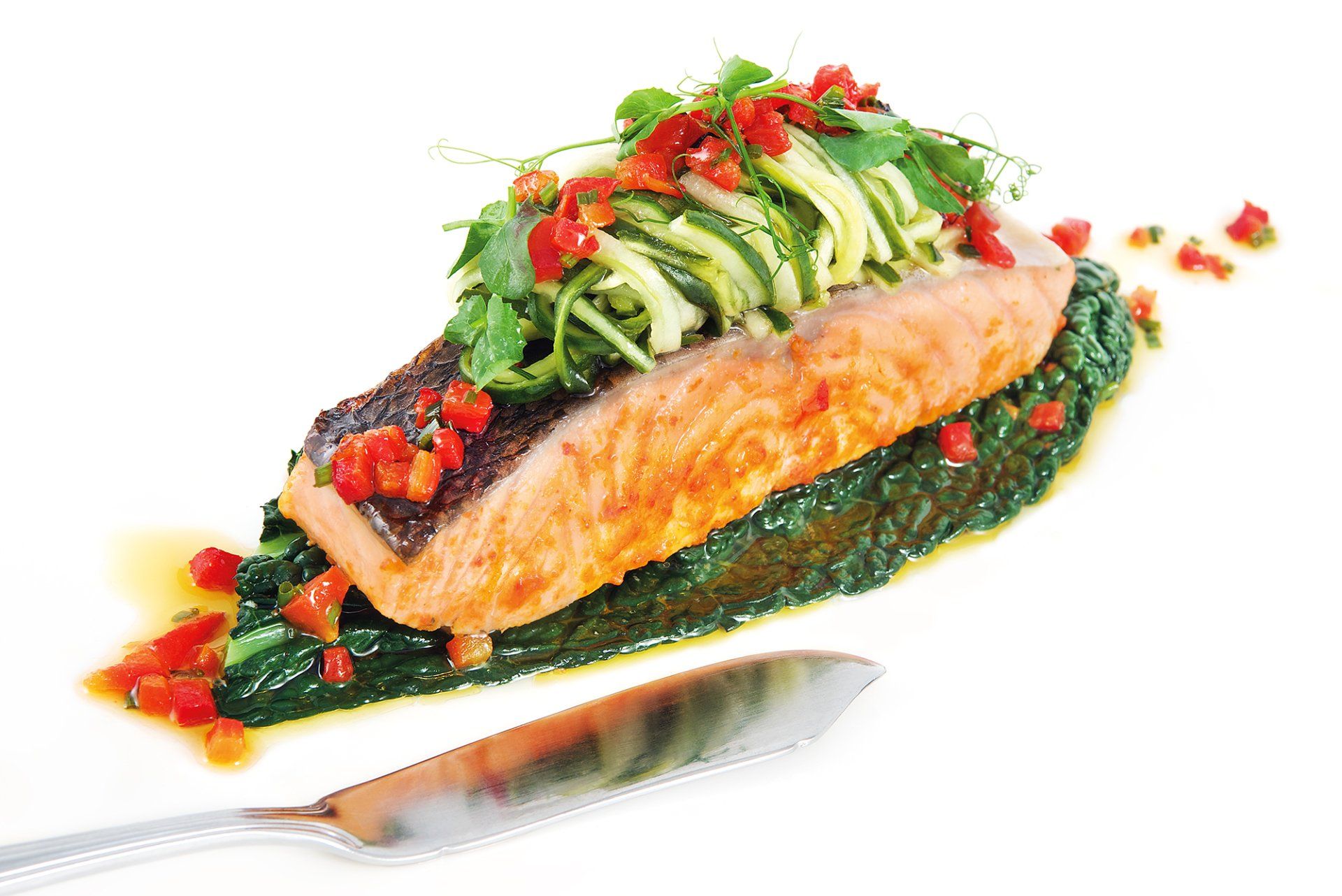 Seared salmon fillet with green vegetables, red peppers, and greens on white plate with knife.