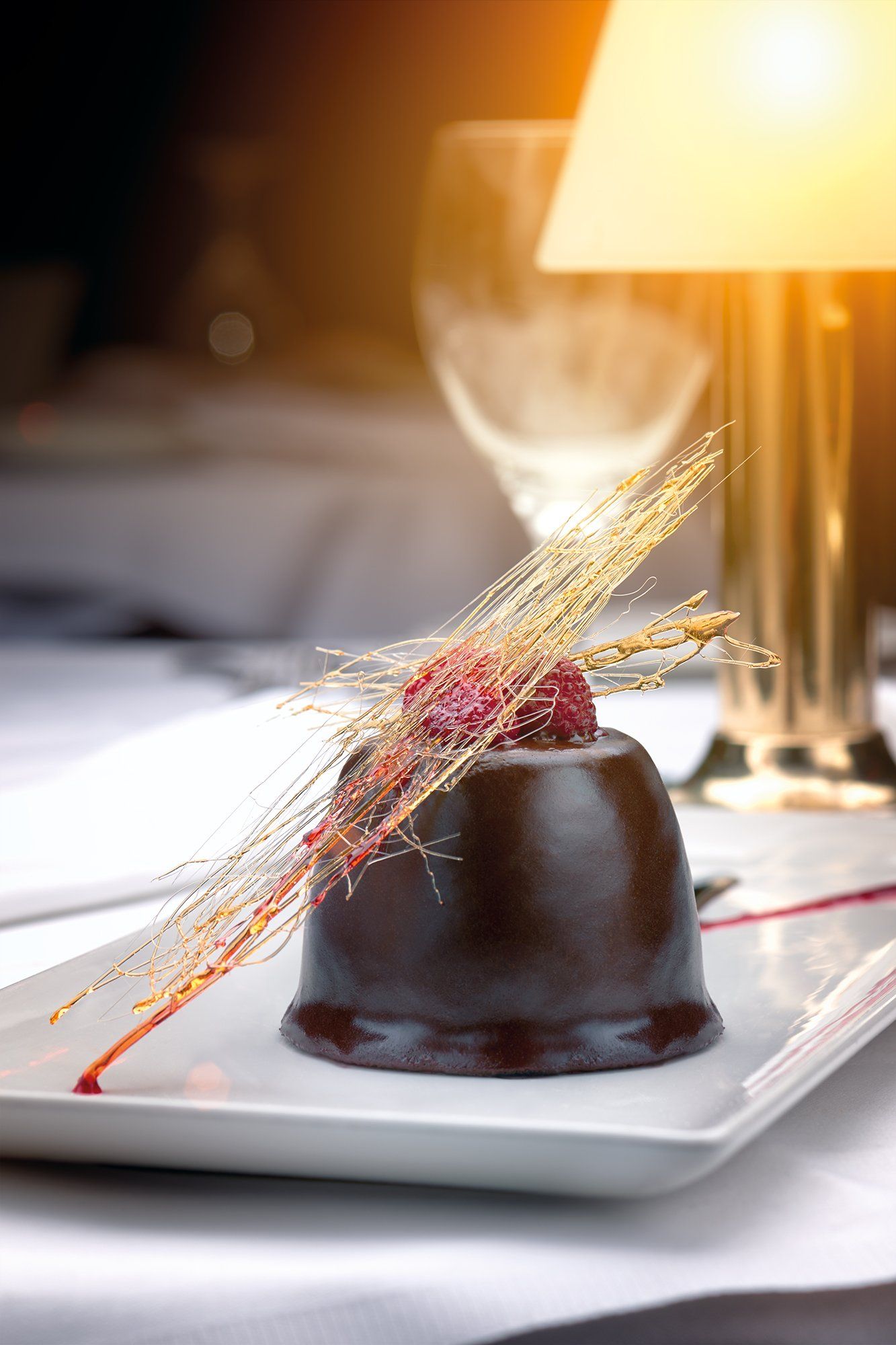 Chocolate dessert with raspberries and spun sugar on a white plate, set on a table with a lamp.