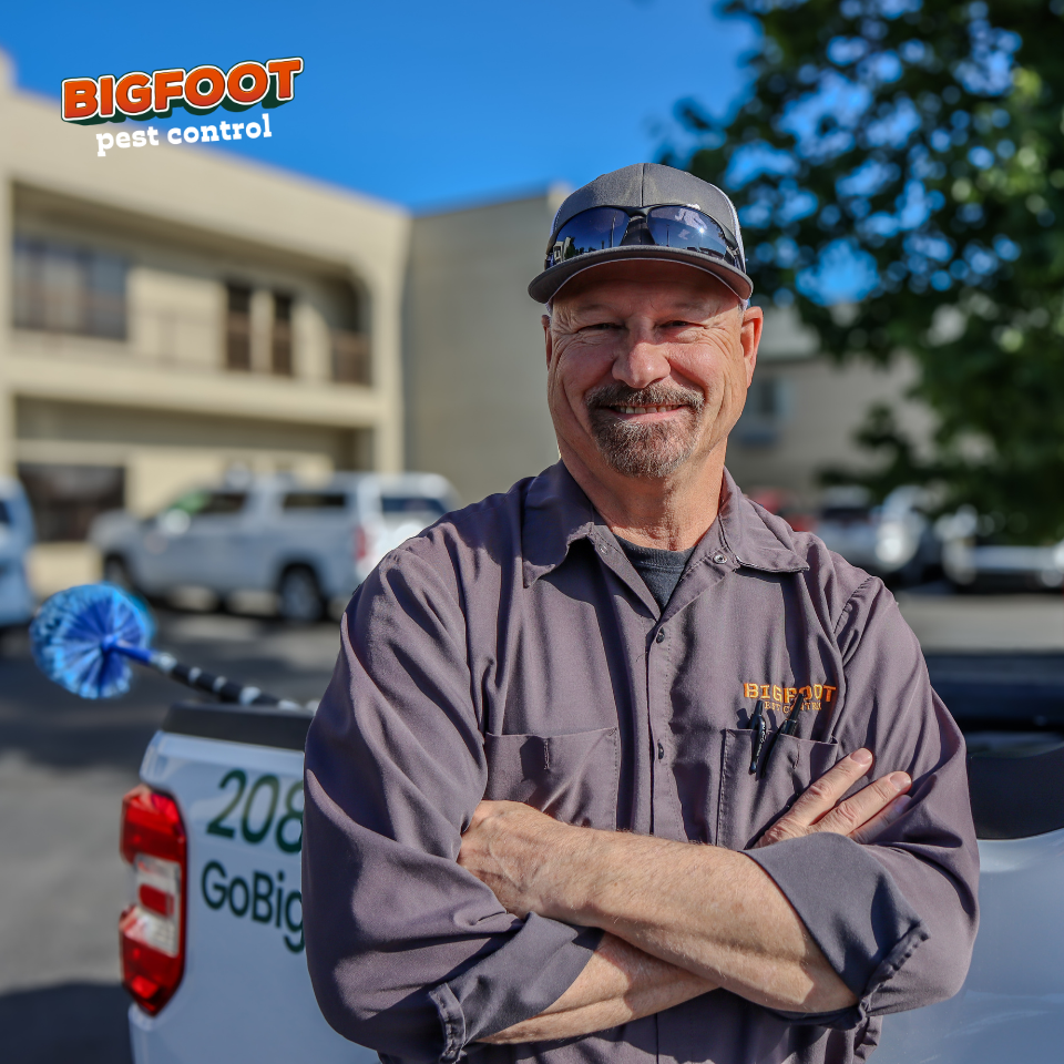 A Bigfoot pest control technician wearing a hat and glasses is standing in front of a truck.