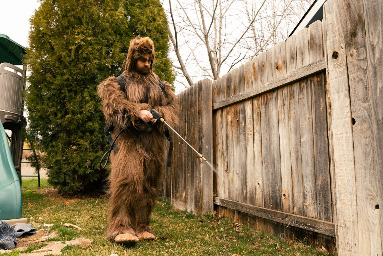 A man in a bigfoot costume is spraying a wooden fence with a hose.