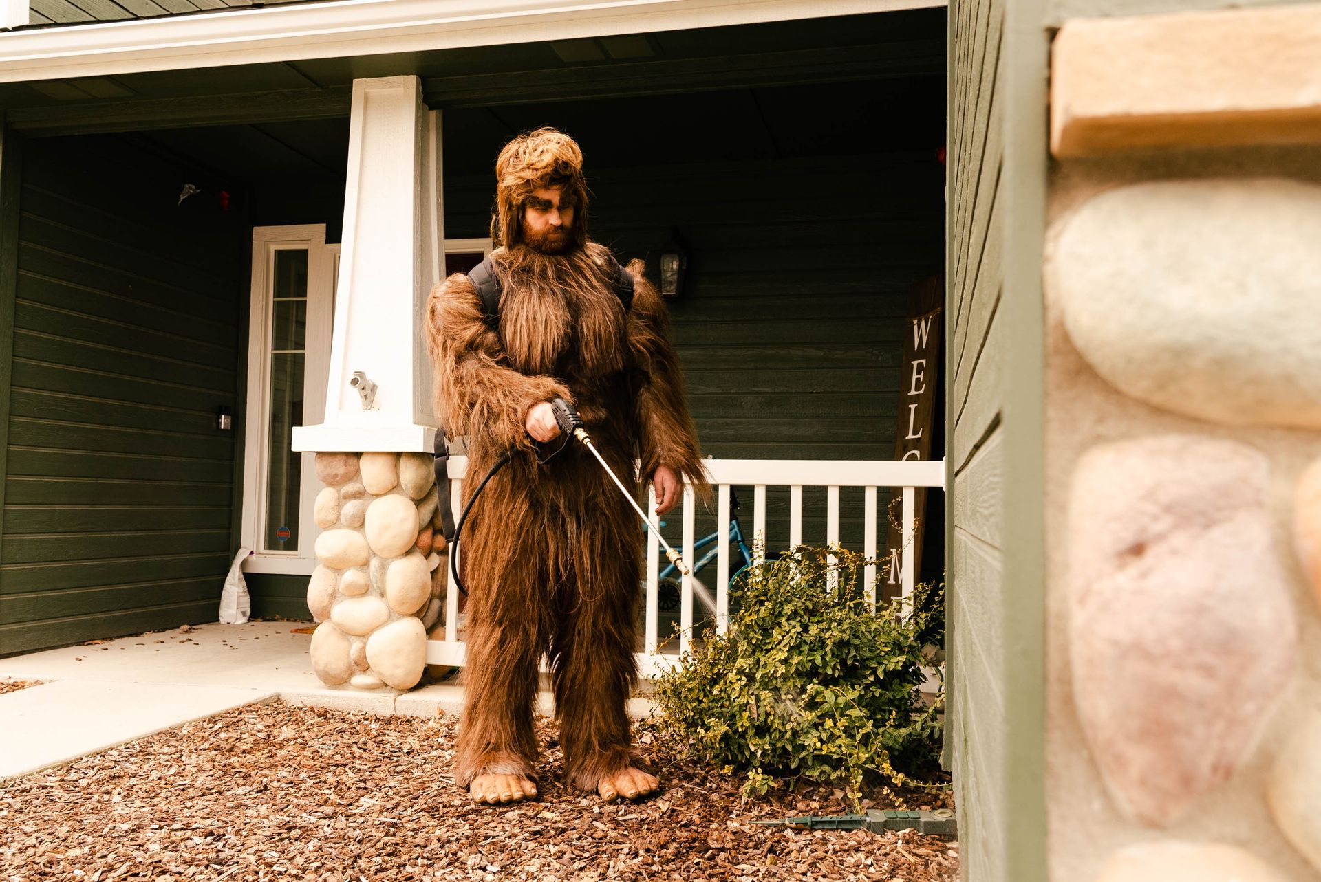 A man in a bigfoot costume is standing in front of a house, spraying for pests