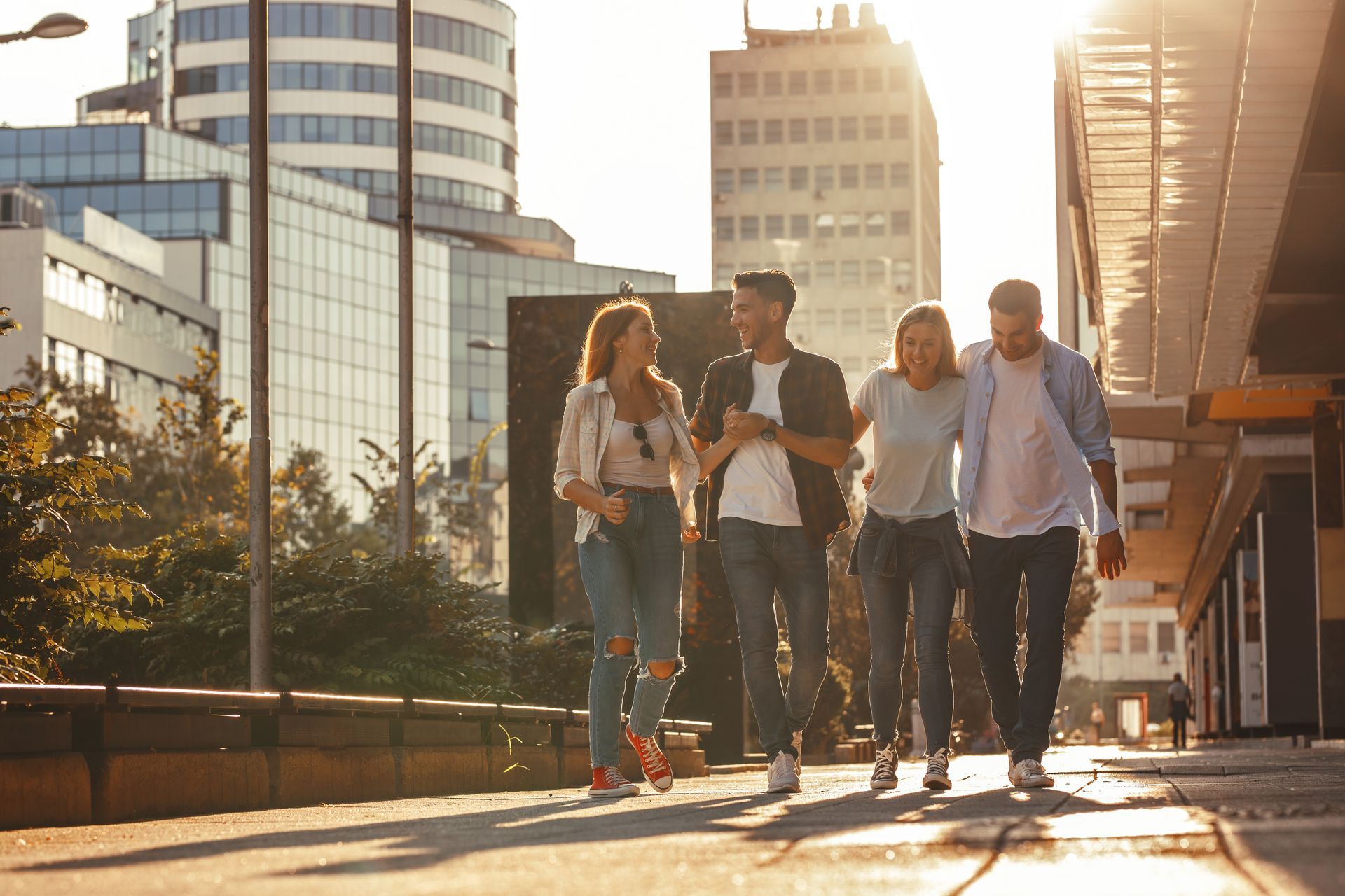 Four young adults walking and talking together down a city sidewalk, sunny day.