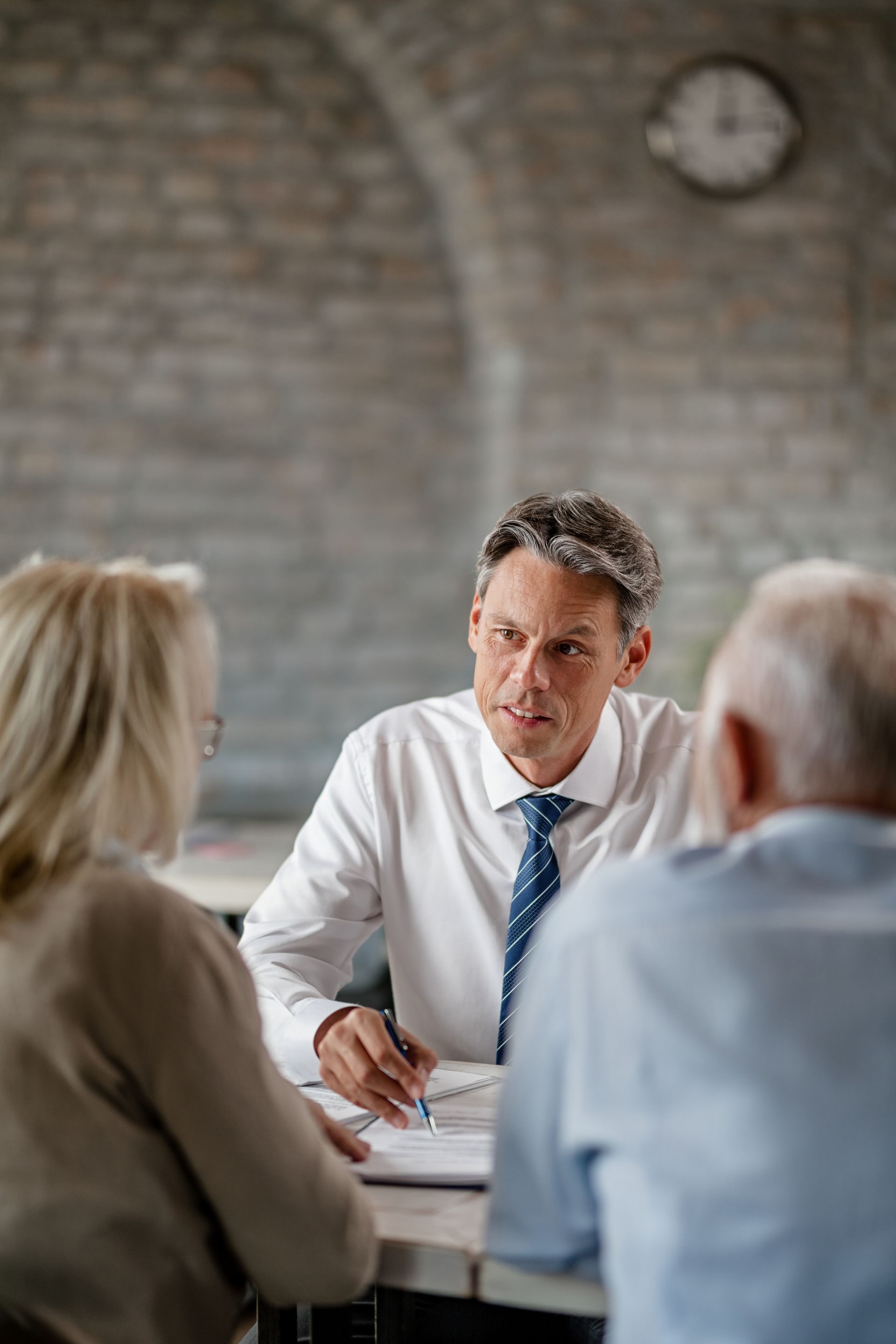 Man in a white shirt and tie consults with an older couple at a table. Brick background.