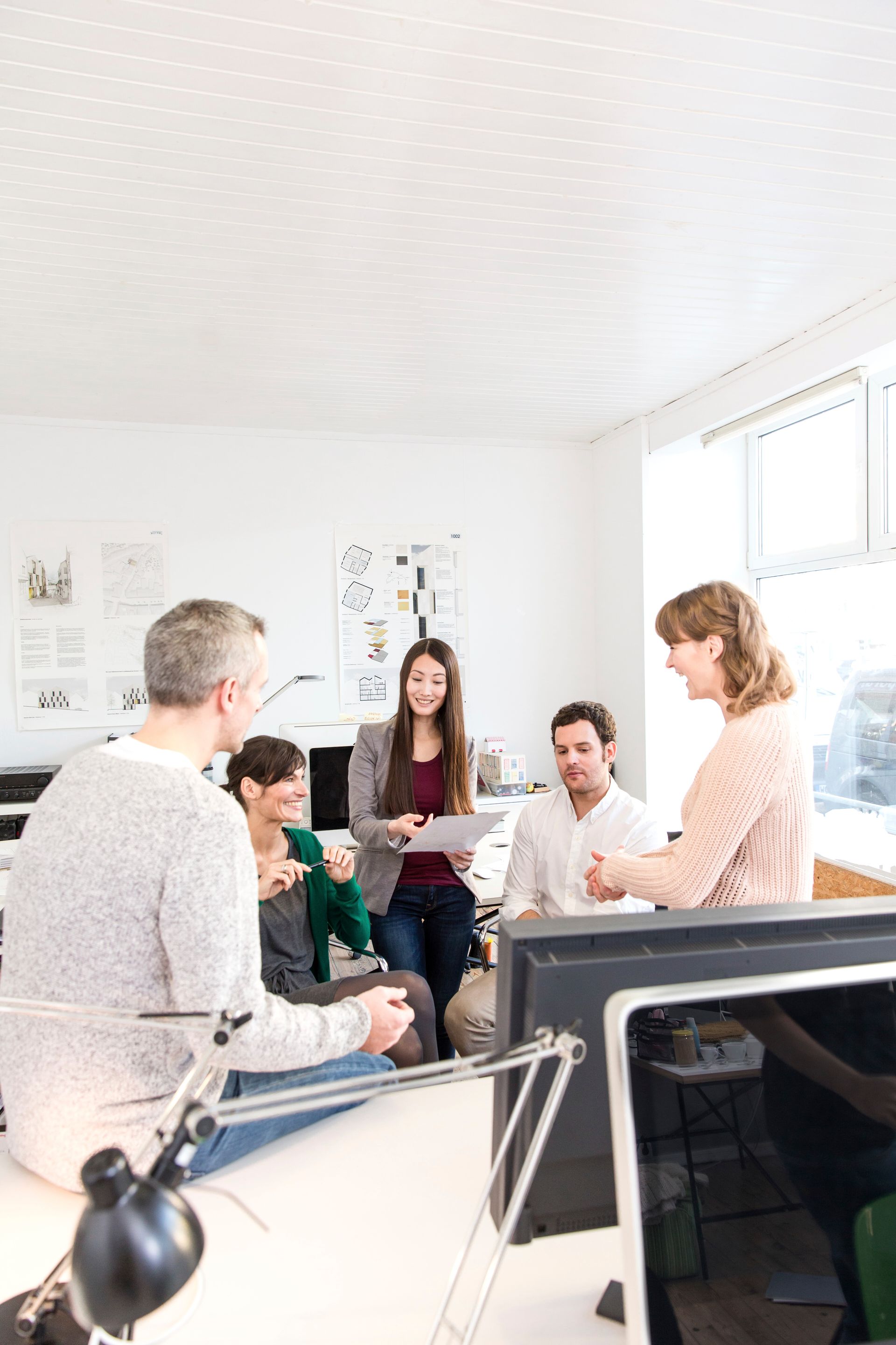 Group of people in a bright office, discussing papers. A woman holds documents, others listen.