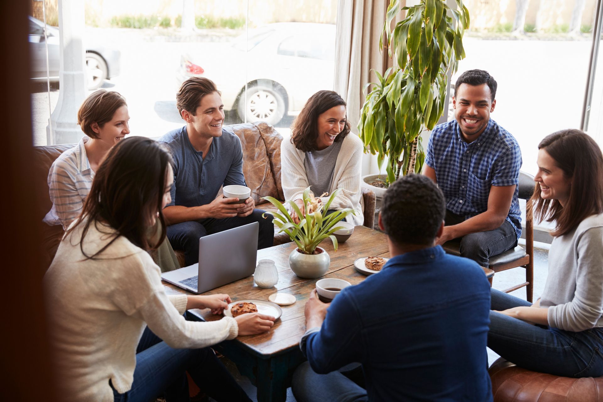 Group of diverse people laughing, gathered around a table with food and a laptop in a bright cafe.
