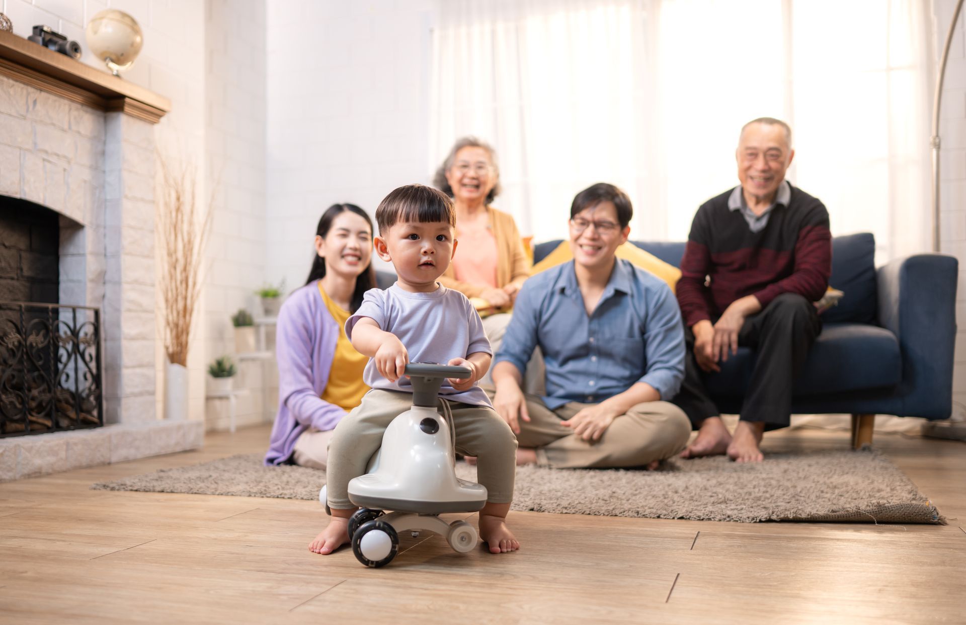 A multi-generational family watches a young child ride a toy car in a living room.