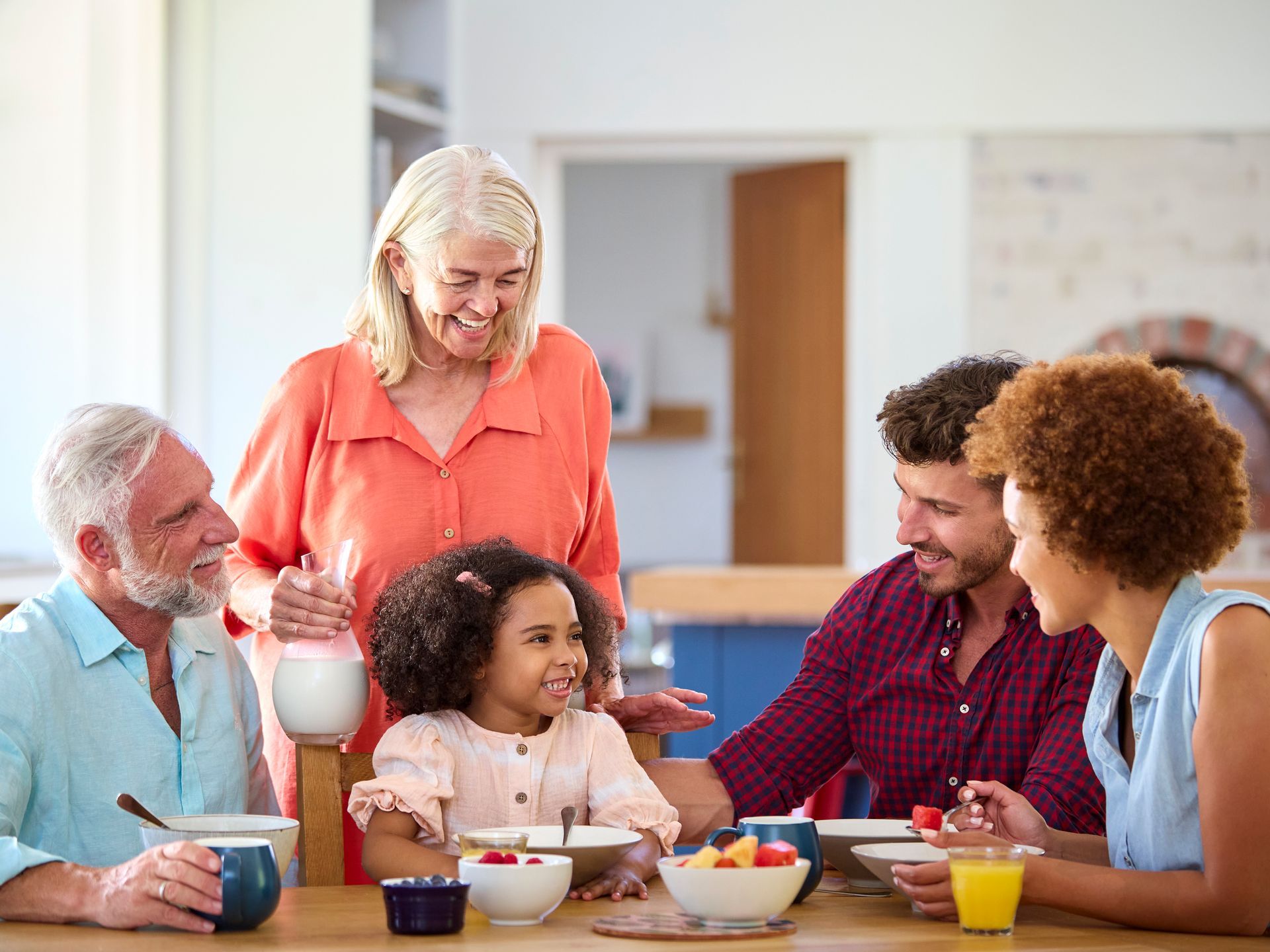 Family gathered around a table, smiling and eating breakfast. Grandmother pouring milk for child.