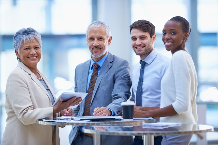 Four diverse professionals smile, gathered around a table indoors, with a tablet and coffee cup.