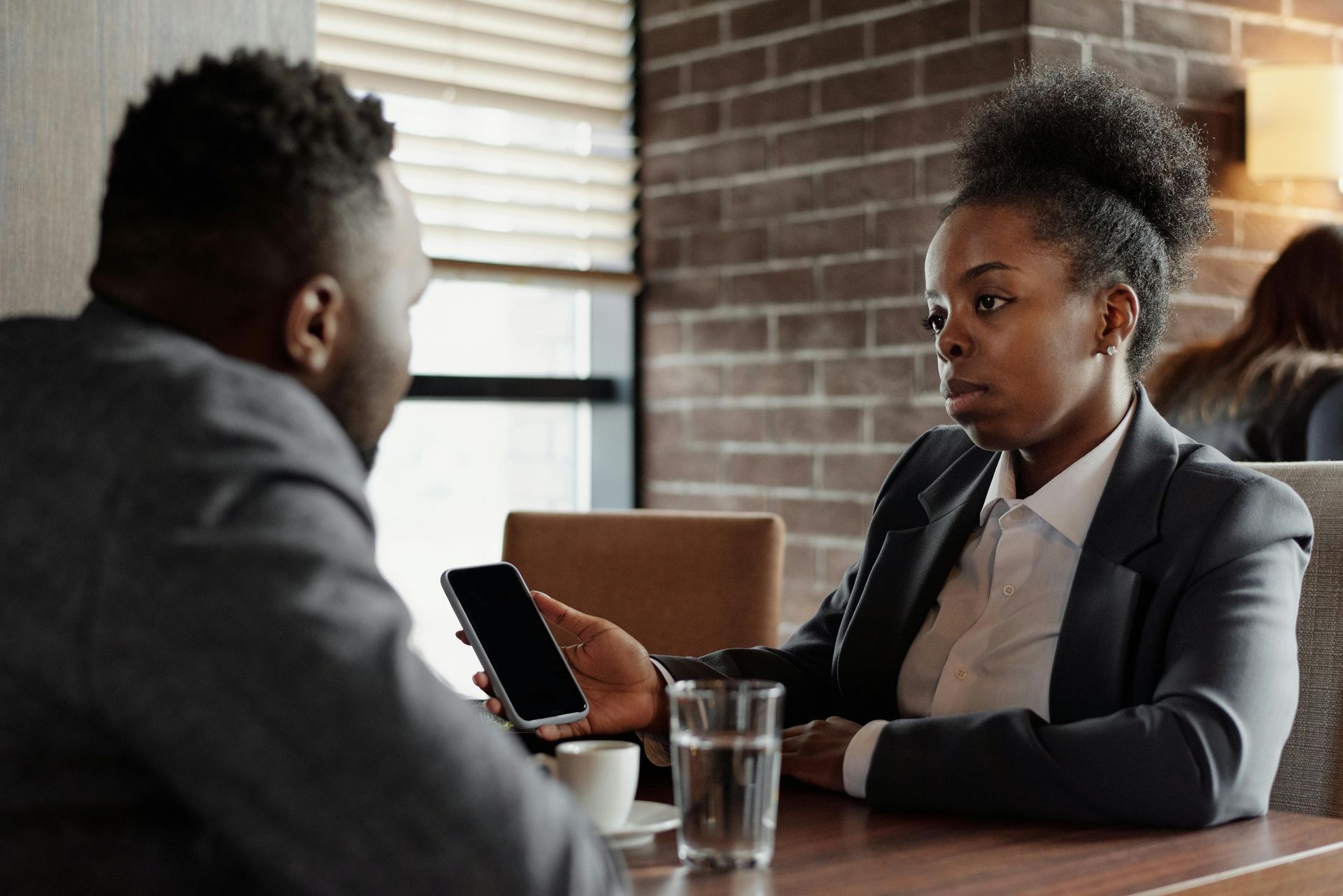 Two businesspeople seated at a table; woman shows man her phone. Brick wall in background.