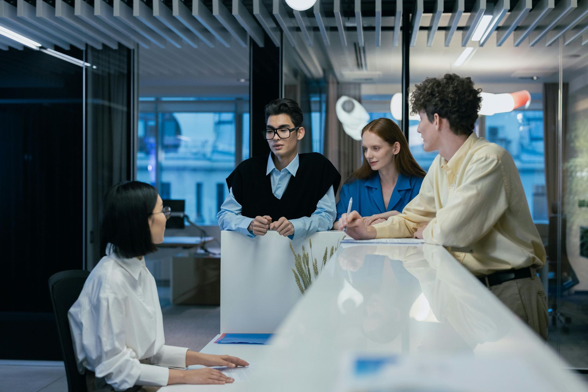 Four people at a reception desk discussing documents, a woman seated, three standing. Modern office.