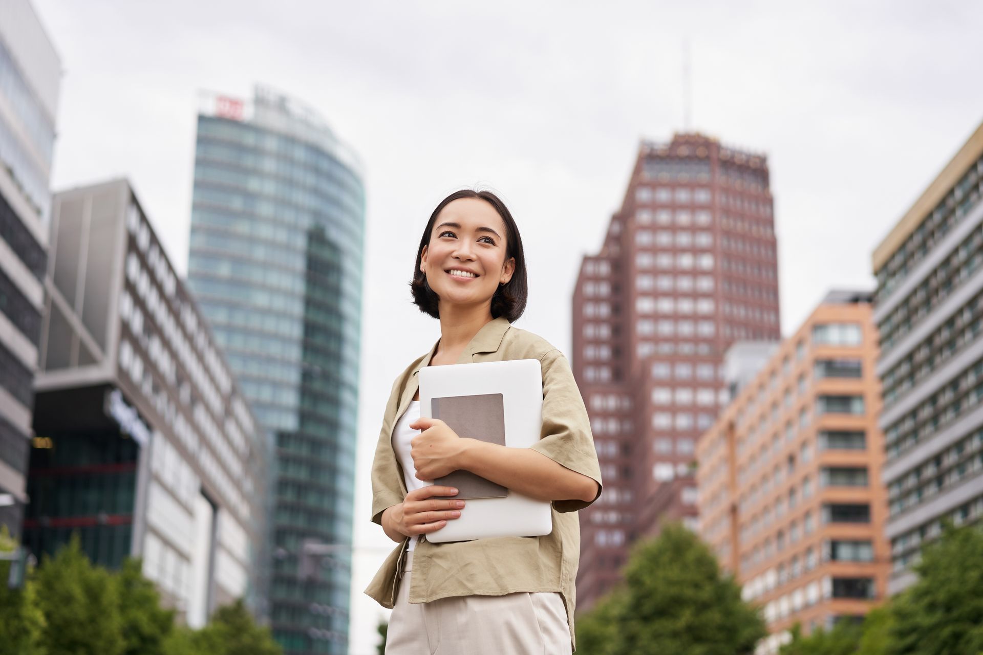 Woman smiling, holding laptop, standing on a city street with tall buildings.