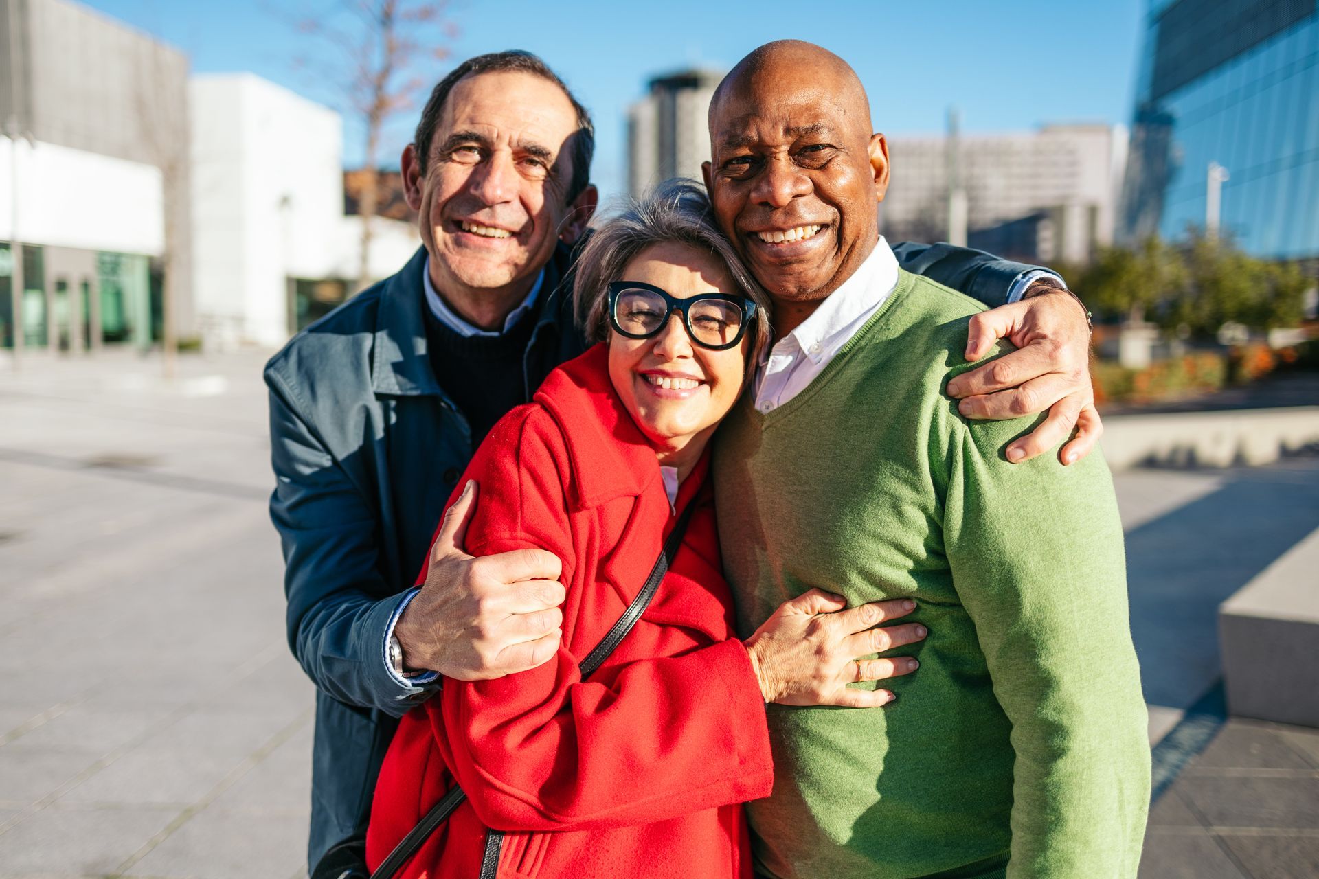 Three smiling friends, diverse, embracing outdoors. Woman in red coat, man in green, other man in jacket. Sunny day.