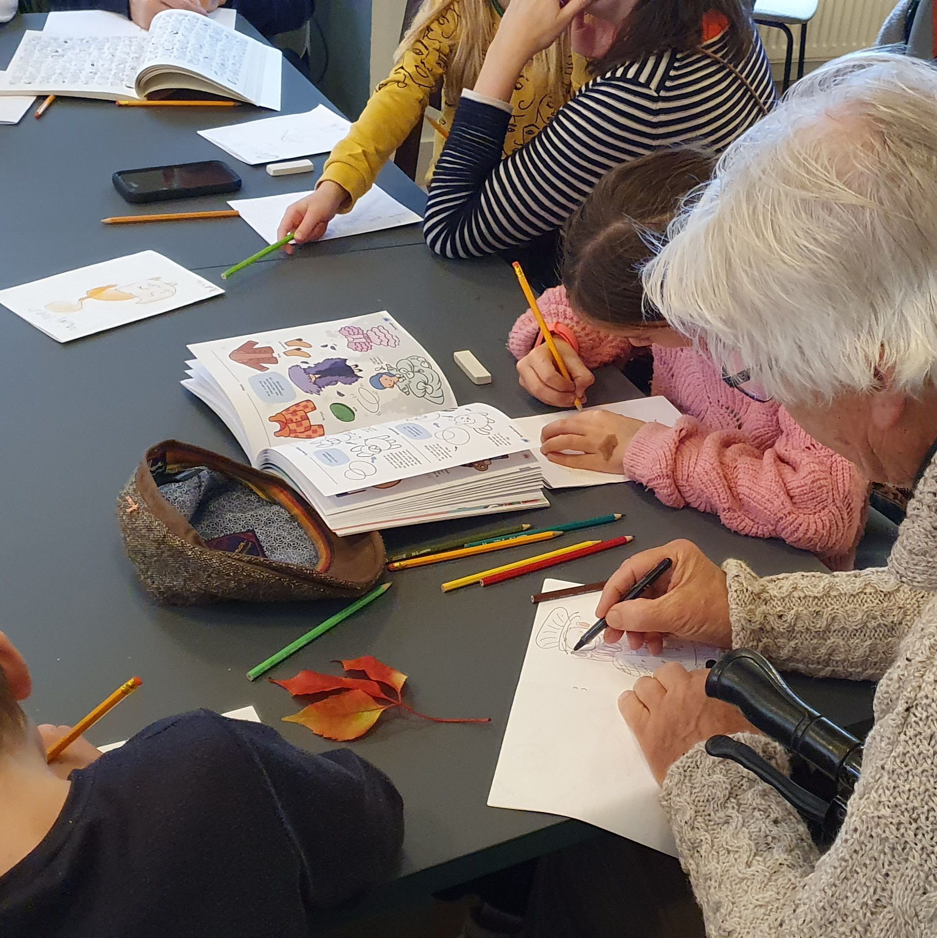 Mensen zitten aan een tafel te tekenen met boeken en potloden. Eén van hen gebruikt een wandelstok.