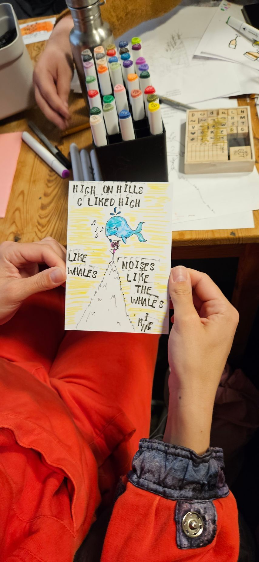 Een persoon houdt een tekening van een blauwe walvis boven een berg vast, met tekst erbij. Op de achtergrond zijn kleurrijke stiften en tekenbenodigdheden te zien.