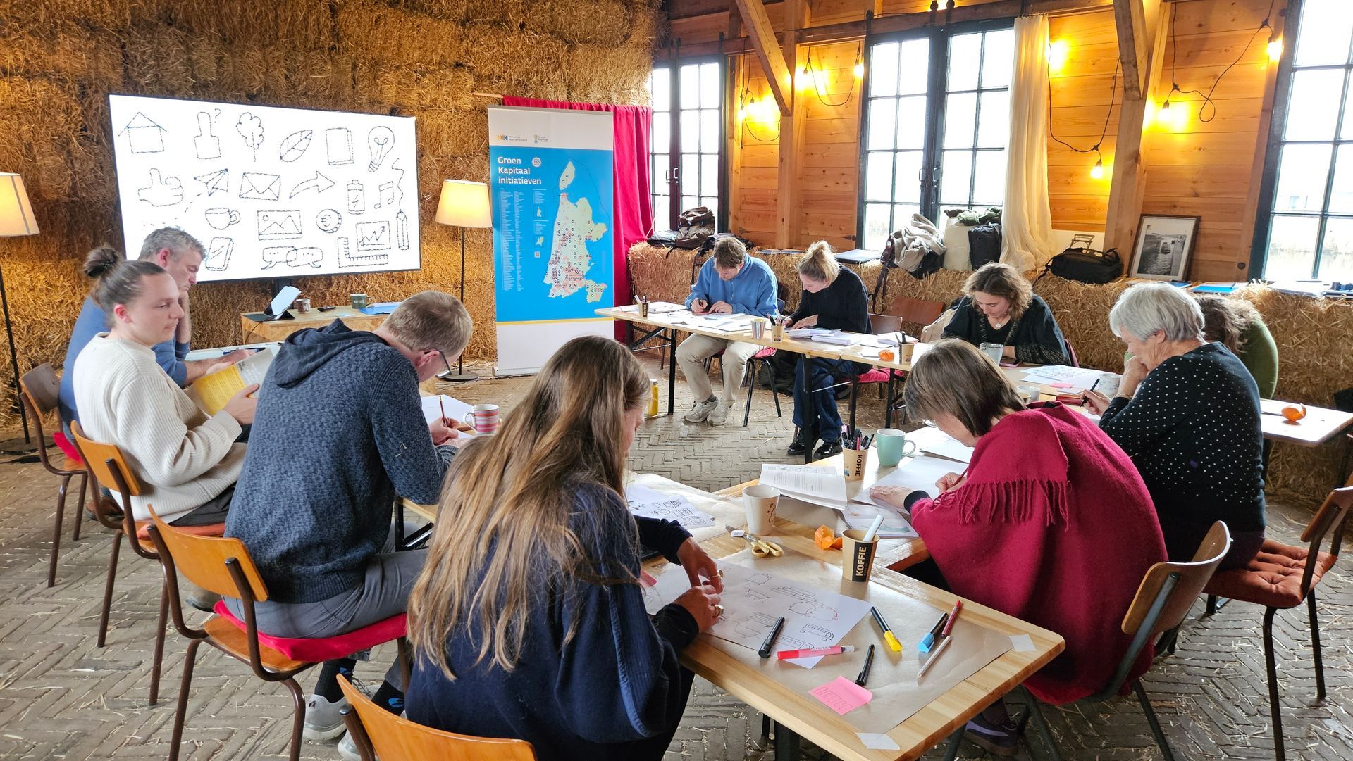Mensen zitten aan tafels in een rustiek gebouw en werken aan projecten. Een spandoek en een whiteboard zijn zichtbaar.