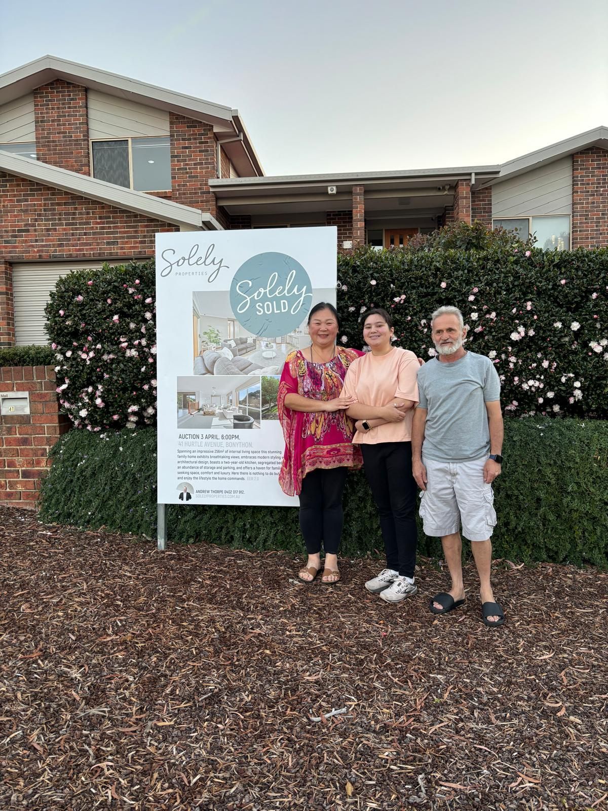 Three people stand by a white sign in front of a brick building and hedges.