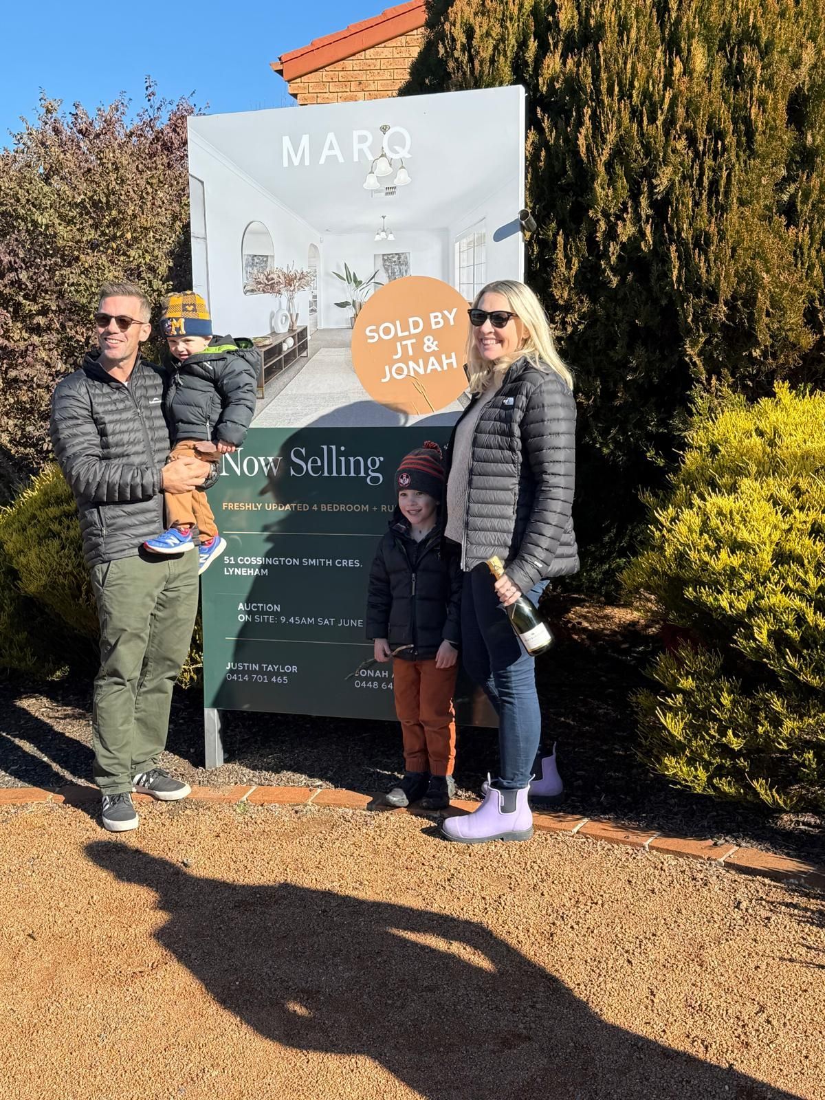 Family posing beside a real estate sign outdoors on a sunny day