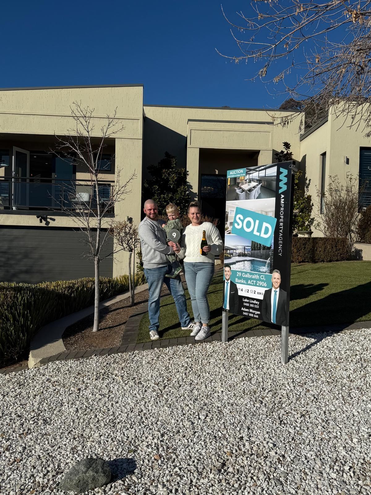 Three people posing beside a real estate sold sign outside a tan building on a sunny day