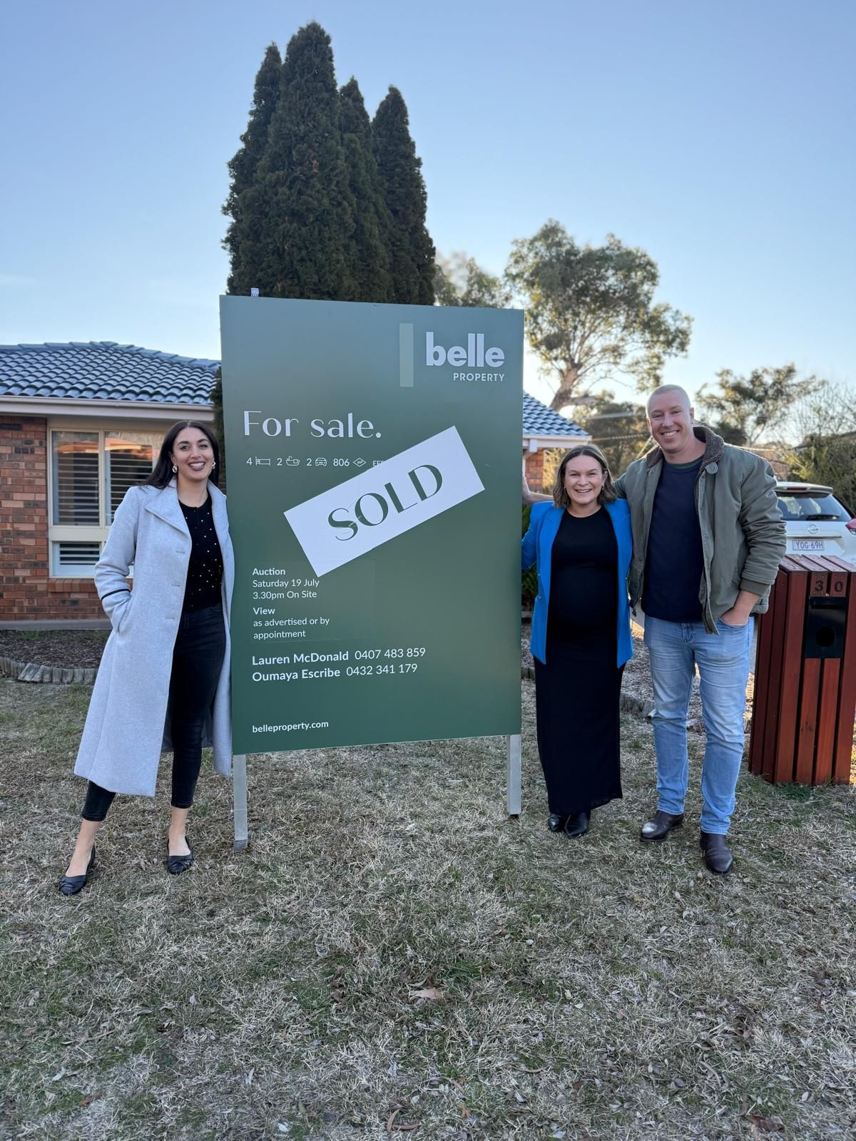 Three people posing beside a sold real estate sign in front of a house.
