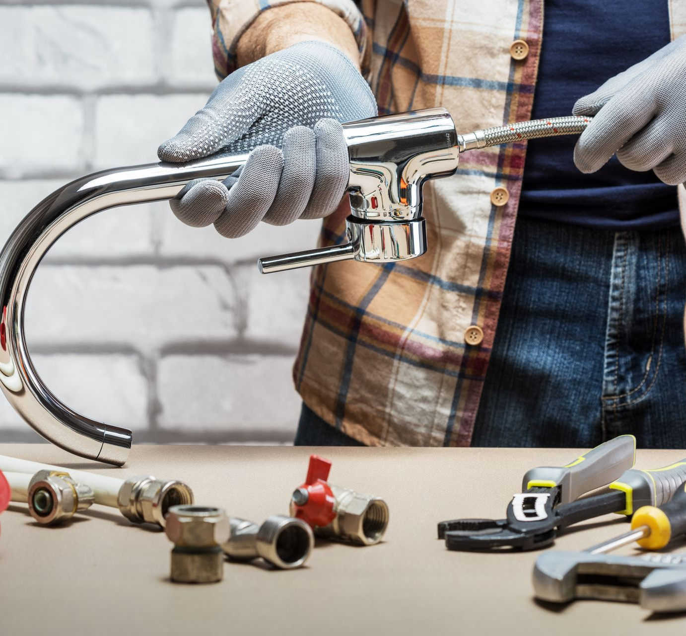 A person wearing work gloves holds a chrome kitchen faucet above a surface with plumbing tools and various pipe fittings.
