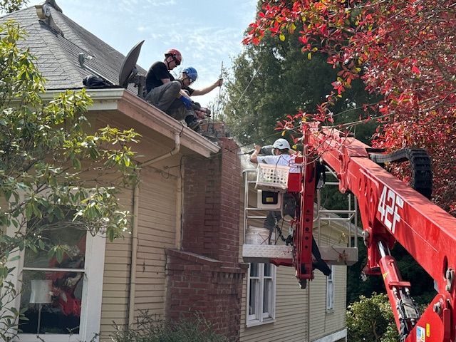 Three people on a roof, one in a lift, working on a house, presumably a chimney. Red leaves and blue sky in the background.