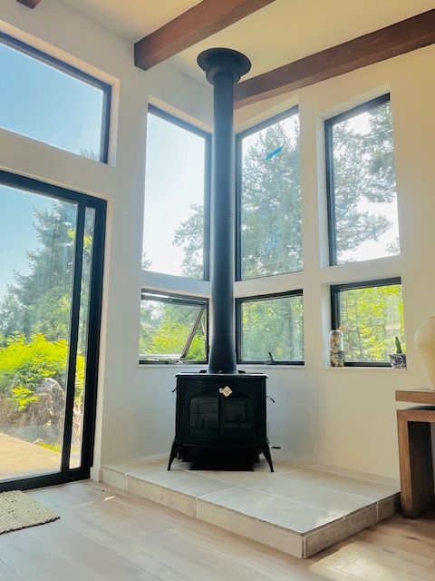 Black wood-burning stove with tall chimney against a backdrop of large windows.  Sunlight streams through the windows, illuminating the white-walled interior.