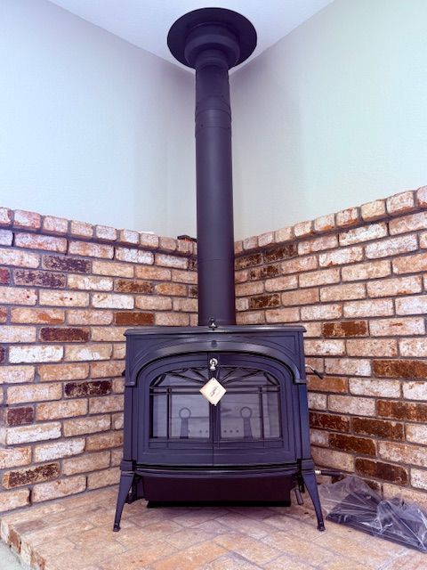 Black wood-burning stove with chimney against a brick wall in a corner of a room. Light-colored walls surround.