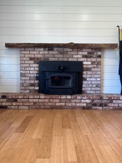 Fireplace with red brick surround and wooden mantel, set against white shiplap. Light wood flooring in the foreground.